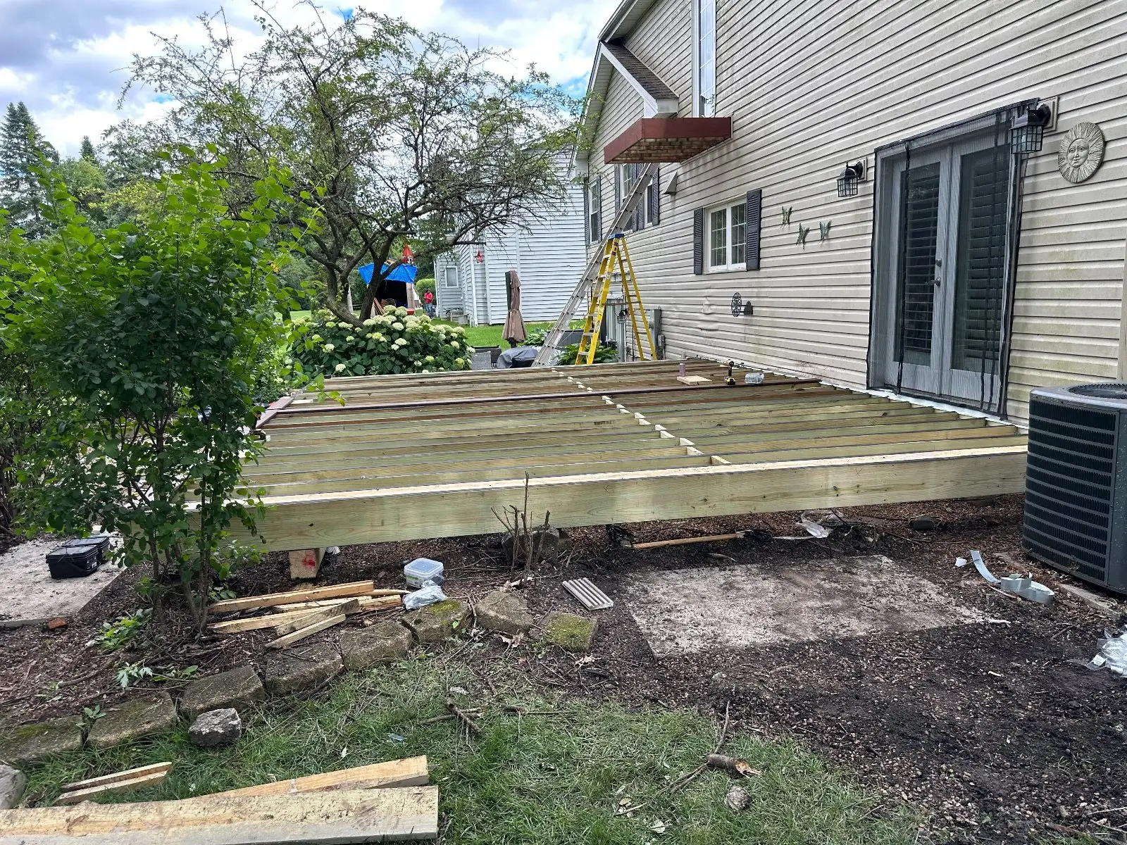 The full new deck frame from the yard, ladder against the house, before the boards go on