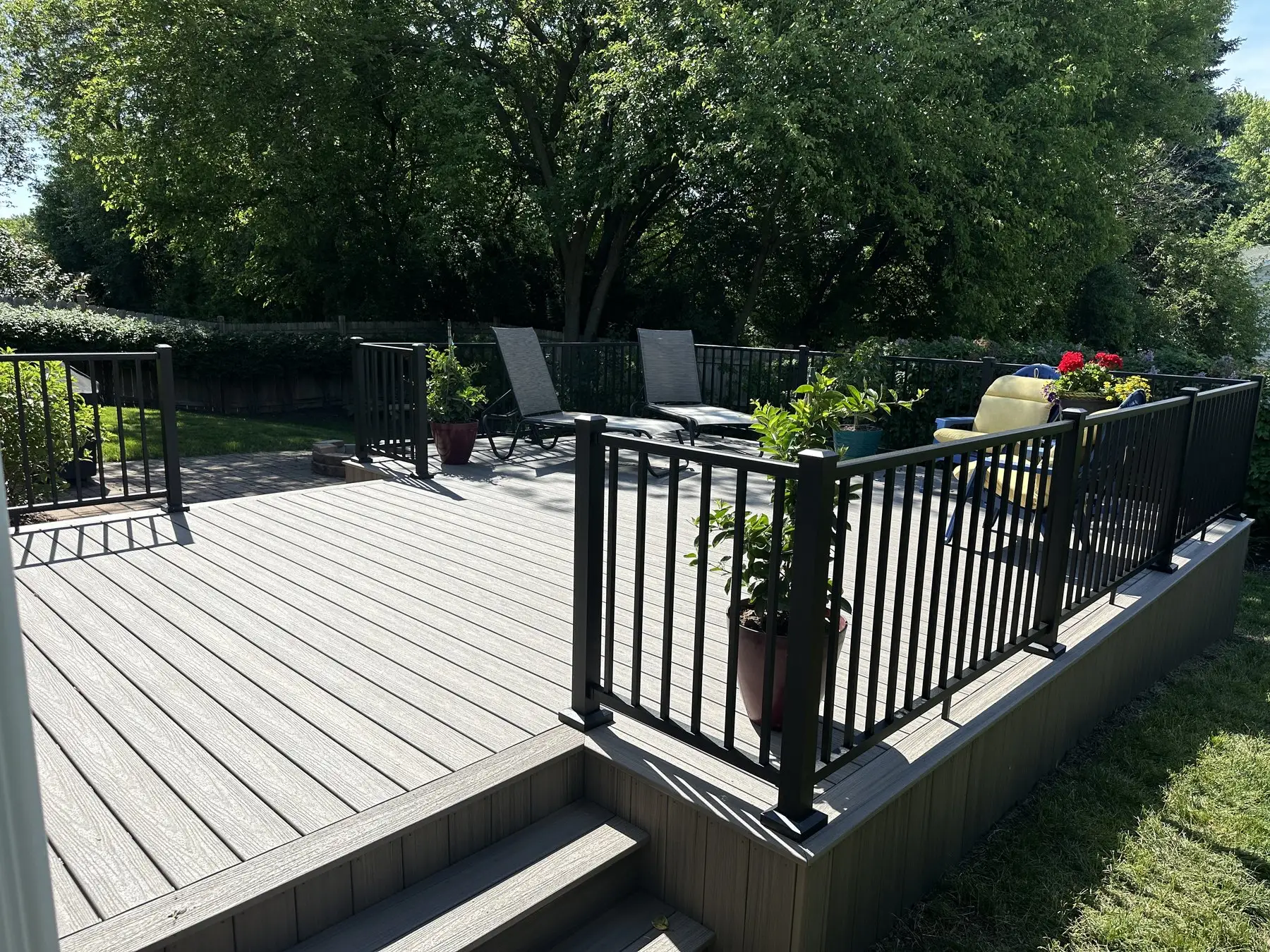 Grey composite deck with black aluminum railings and a wide step down to a paver patio with lounge chairs