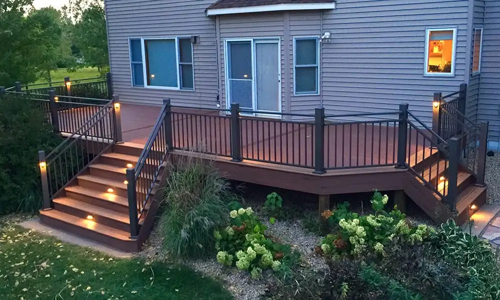 Composite deck lit at dusk, with black railings, warm lighting on the steps and planting beds along the base
