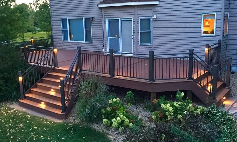 Composite deck lit at dusk, with black railings, warm lighting on the steps and planting beds along the base