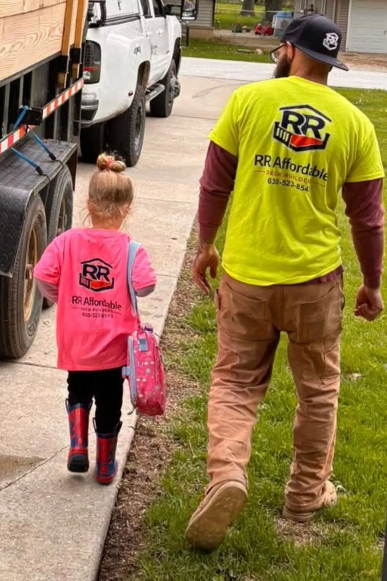 Ryan Miser and his young daughter walking up a driveway to a job site, both wearing RR Affordable Deck Builders shirts, past a truck and a trailer loaded with lumber